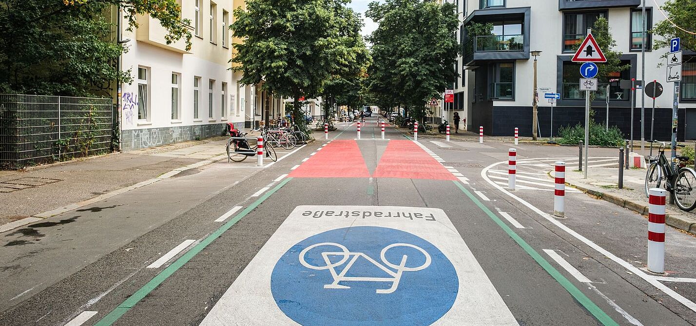 Fahrradstraße Berlin Eine Straße in Berlin: Rechts und links sind Häuserzeilen zu sehen, die Fahrbahn ist mit einem großen Fahrradsymbol versehen. Weiter hinten ist die Fahrbahn rot eingefärbt, Poller dienen als modaler Filter.